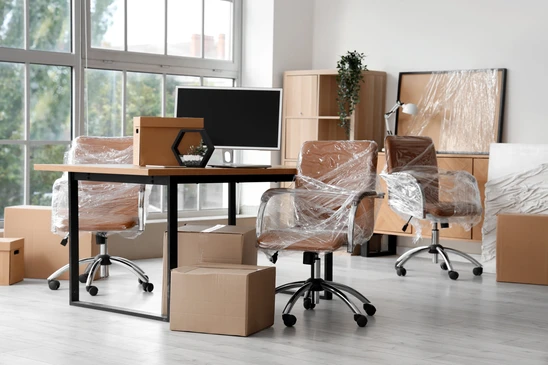 Bureau d'entreprise en cours de déménagement avec chaises de direction en cuir marron emballées sous film plastique, cartons de déménagement, mobilier de rangement en bois clair, écran d'ordinateur sur table noire